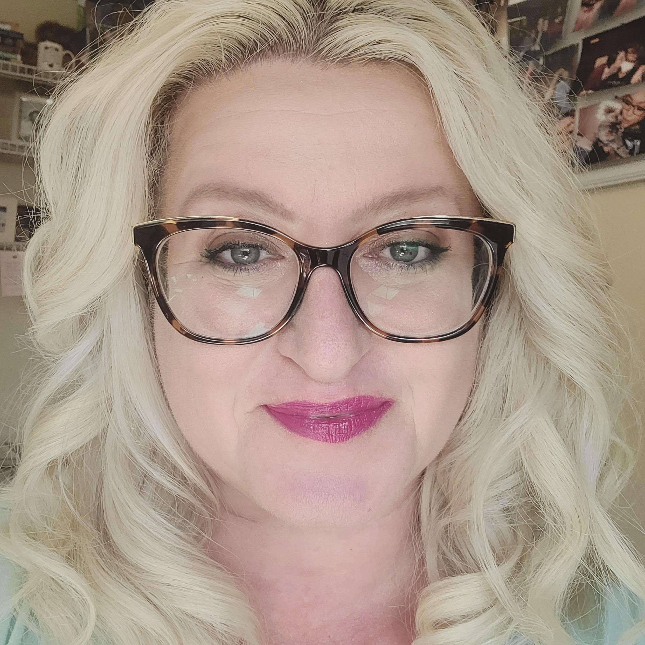 A portrait of Reny with blonde, wavy hair and stylish glasses, smiling warmly. She is in a cozy, personal space with a soft, inviting ambiance.
