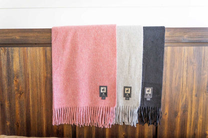 Alpaca wool throw blankets in pink, gray, and black colors with fringe, displayed on a wooden surface.