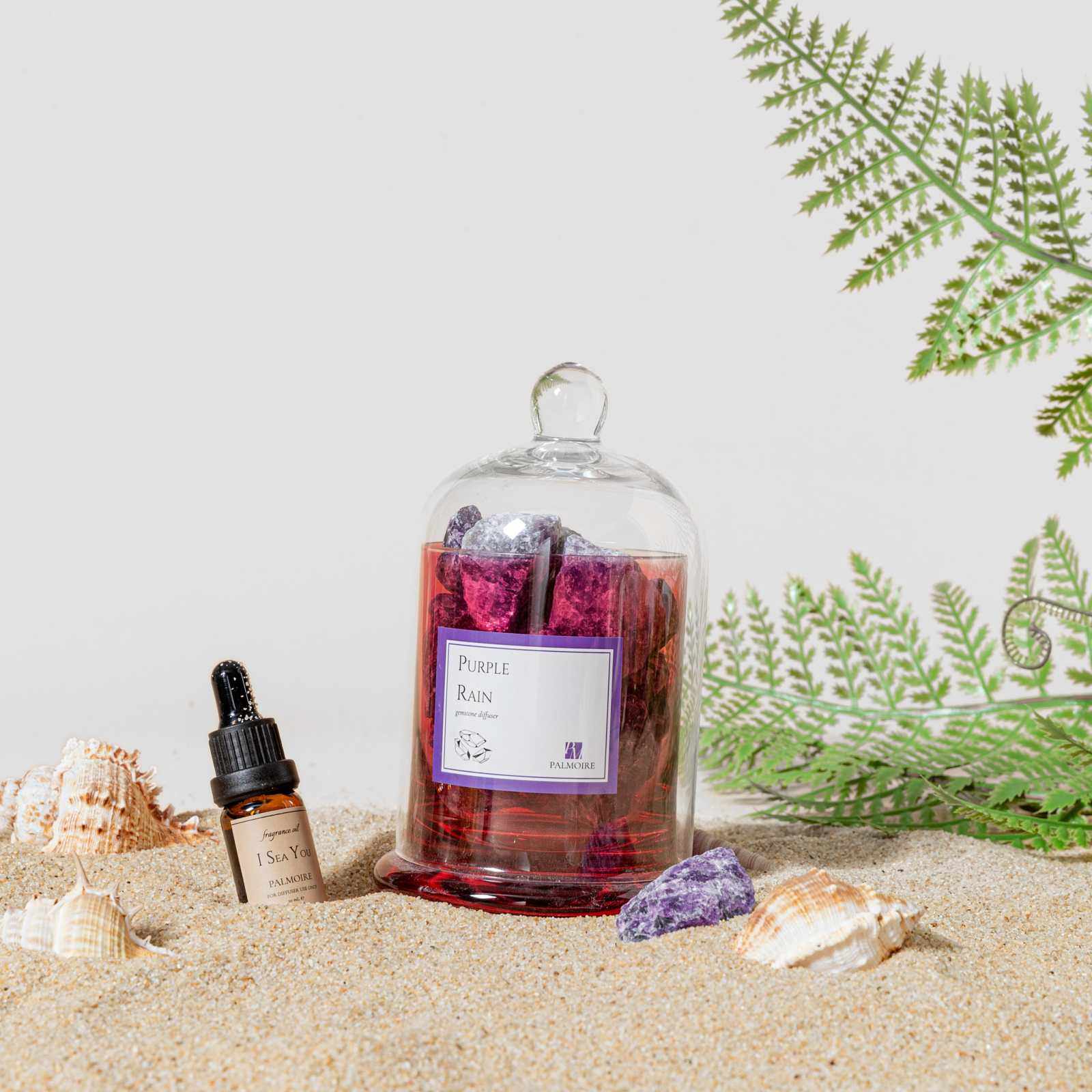 Purple Rain Gemstone Diffuser by PALMOIRE with I Sea You Fragrance Oil on sandy beach with shells and greenery.